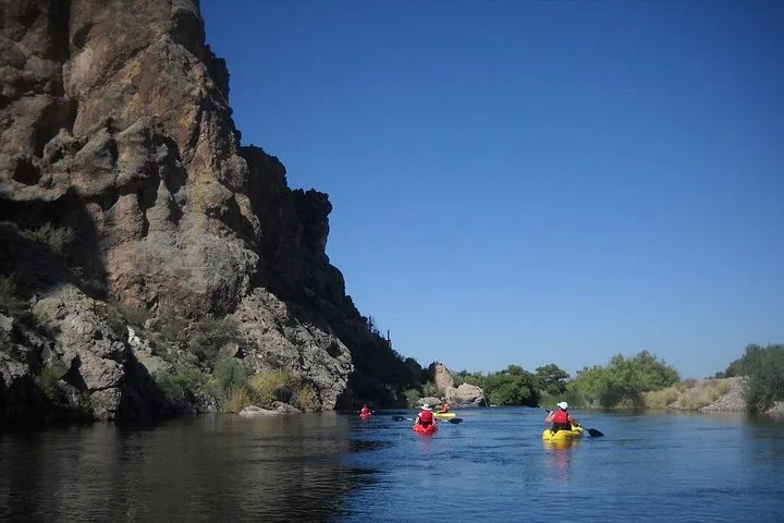 Kayakers paddle on a calm river beside a large rocky cliff